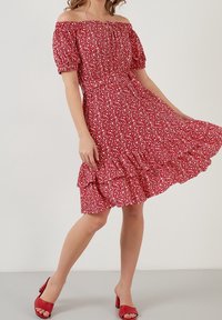 Femme portant une robe fleurie rouge à épaules dénudées avec des manches bouffantes, tenant sa jupe tout en se tenant debout en sandales à talons hauts rouges sur un fond uni.