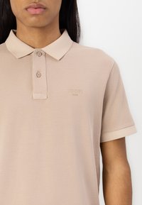 Béžové polo tričko vyrobené z texturované tkaniny, s klasickým límcem, dvoubarevnou knoflíkovou légou a decentním embosovaným logem na hrudi.