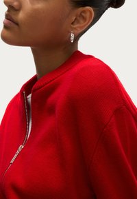Maglione rosso con zip, colletto a costine e texture lavorata a maglia, abbinato a orecchini a cerchio argentati con un design colorato.