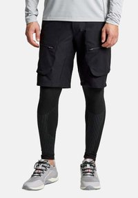 Pantaloni cargo neri con tasche con zip, abbinati a reggiseni neri testurizzati con strisce verticali e scarpe da ginnastica grigie.