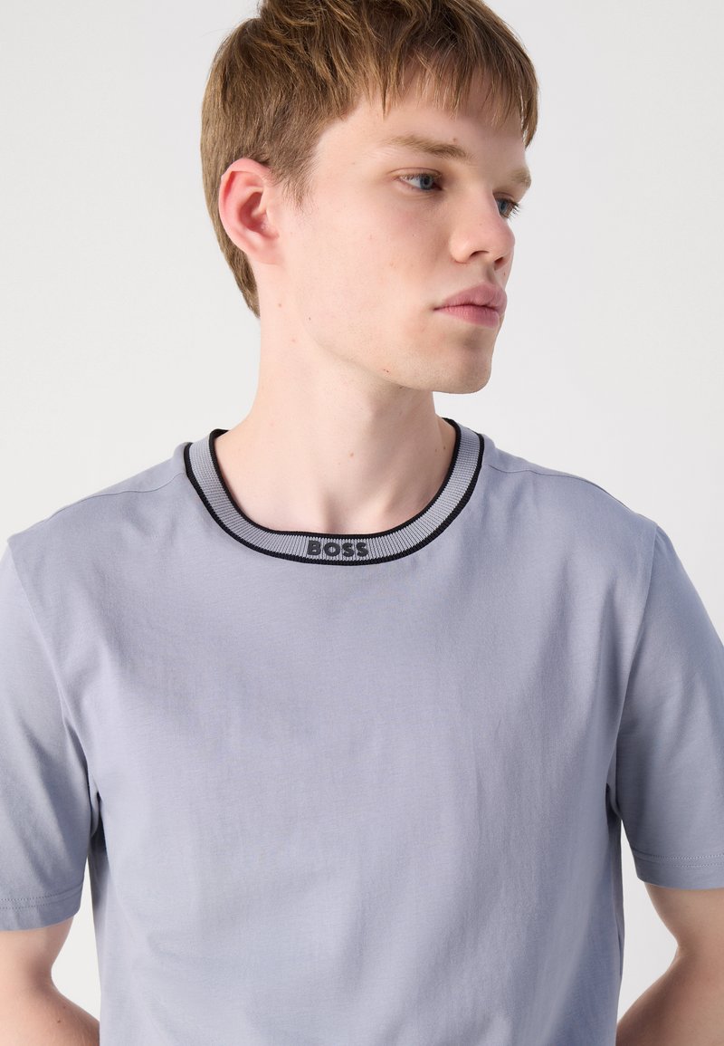 T-shirt grigio chiaro con maniche corte, colletto a costine nero e grigio e una texture sottile. Logo visibile sul colletto.