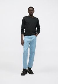 Čierna mikina s dlhým rukávom, svetlomodré rovné džínsy a čierne robustné čižmy. Model stojí pred jednoduchým svetlým pozadím.