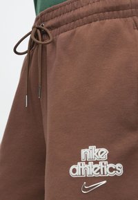 Καφέ βαμβακερά σορτς τζόγκινγκ με ρυθμιζόμενη μέση με κορδόνι, με λευκό λογότυπο «nike athletics» και μεταλλική λεπτομέρεια στις άκρες του κορδονιού.