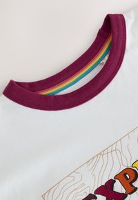 Fehér pamut póló bordó kerek nyakkivágással. Színes csíkos díszítés található a gallér belsejében, valamint mintás nyomat az elején.