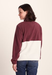 Woman with shoulder-length dark hair wearing a two-tone fleece jacket in maroon and white, paired with blue jeans, facing away to the left.