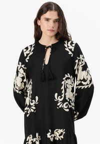 Sort bluse med beige barok mønster, nøglehulshalsudskæring med frynser, lange ærmer og en afslappet pasform. Glat stoftekstur.