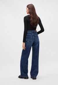 Široké džínsy v tmavomodrom denime, s vysokým pásom, dvoma zadnými vreckami a koženou nášivkou na zadnej strane. Kombinované s čiernym topom.