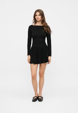 Νέα γυναίκα με μακριά καστανά μαλλιά που φοράει μαύρο φόρεμα με μακριά μανίκια και μαύρα παπούτσια Mary Jane, στέκεται μπροστά σε λευκό φόντο.