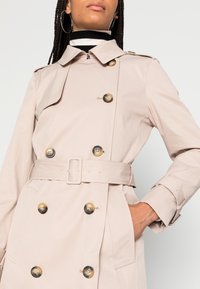 Beige trenchcoat med skärp i midjan, dubbelknäppt fram, stora mörka knappar och en klassisk krage. Stylad med en svartvit randig polotröja.