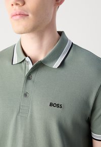 Žalias polo marškinėliai su tekstūruotu audiniu. Yra klasikinis apykaklė su juodai baltais juostelėmis ir mažas siuvinėtas "BOSS" logotipas.