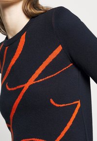 Knit sweater in black with bold red abstract patterns. Features a ribbed crew neckline and long sleeves. Textured fabric details visible.