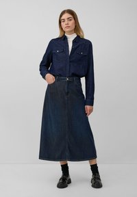 Camicia di denim con due tasche sul petto, colore blu scuro; abbinata a una gonna di denim lunga e svasata. Scarpe nere lucide e calze nere completano il look.
