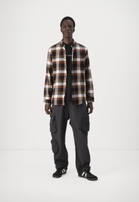 Geverfde shirt in oranje, wit en zwart; zwart t-shirt eronder; donkere cargobroeken met zakken; zwarte sneakers met witte strepen.
