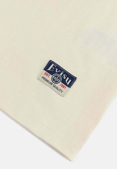 T-shirt di cotone color crema con una toppa rettangolare del logo Evisu con testo blu, "EST. 1991" e "PREMIUM QUALITY." Texture morbida, maniche corte.