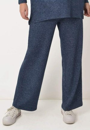 Persoon die losse donkerblauwe broek en witte sneakers draagt, staand met voeten iets uit elkaar op een effen lichte vloer.
