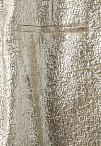 Metalizuoto auksinės spalvos audinio tekstūra, su audiniu raštu ir subtiliomis variacijomis. Įeina maža, diskretiška kišenė.