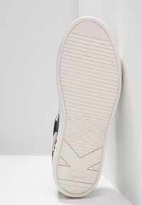 Semelle de chaussure de sport noire avec une bande de roulement en caoutchouc blanc. Présente un motif texturé et le logo gravé "KARL LAGERFELD" sur la semelle extérieure.
