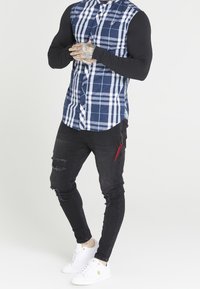 Camicia a maniche lunghe blu e bianca a quadri con maniche nere, abbinata a jeans neri usurati e sneakers bianche con dettagli dorati.