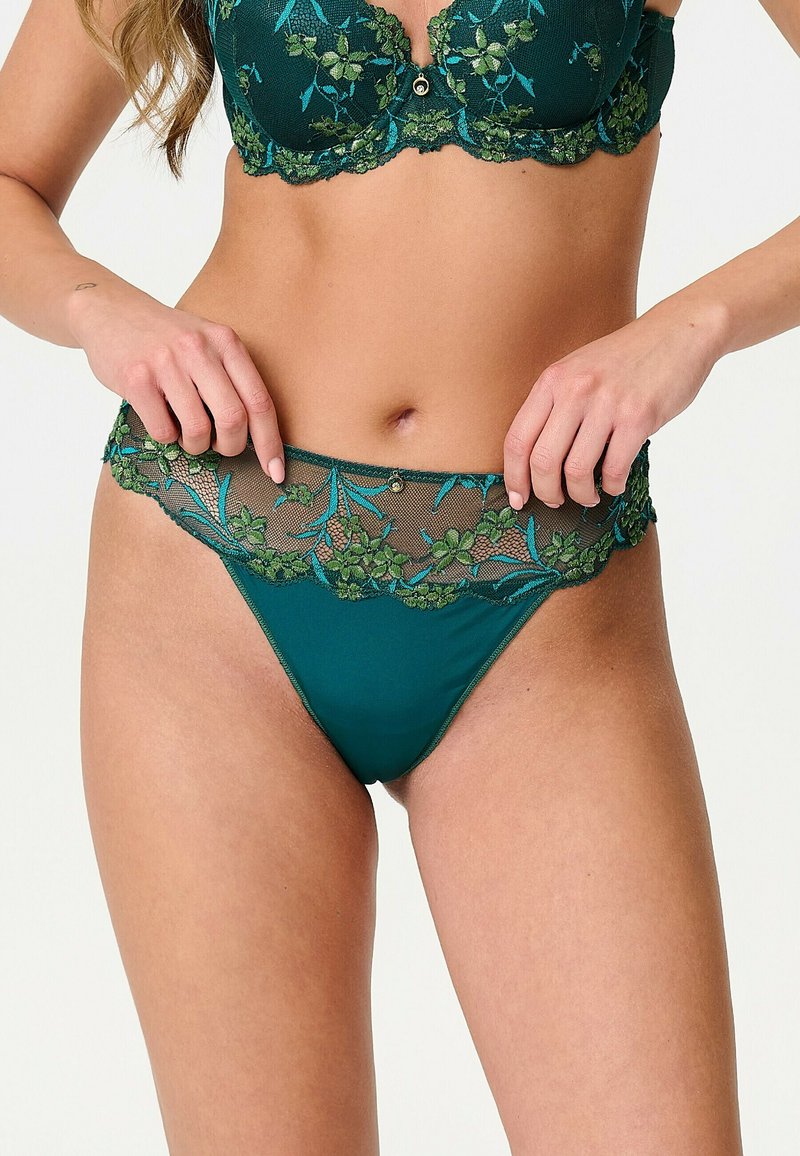 Femme portant un ensemble de lingerie vert foncé avec broderie en dentelle florale et détails en maille transparente, ajustant la taille de la culotte.
