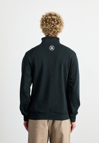 Donkergroene sweatshirt met een hoge kraag, geribde boorden en een cirkelvormig logo op de achterkant. Zachte textuur en een relaxte pasvorm.