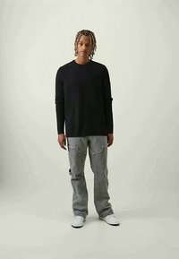 Young man with braided hair stands wearing a black long-sleeve shirt, gray jeans with front pockets, and white sneakers on a plain background.