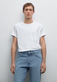 Bílé tričko s krátkým rukávem, s ohrnutými manžetami a kapsičkou na prsou, spárované se světle modrými džíny s rovnými nohavicemi, které mají klasickou sponu a švy.