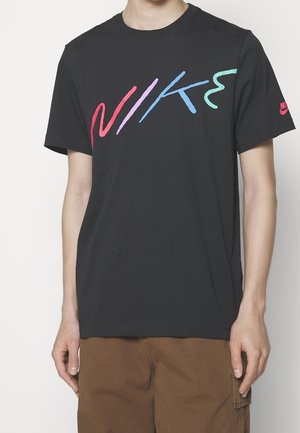 Personne portant un t-shirt noir Nike avec un logo Nike stylisé multicolore sur la poitrine et un logo Nike rouge sur la manche gauche.