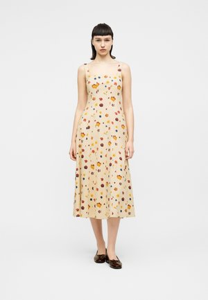 Młoda kobieta stojąca, ubrana w beżową, bezrękawnikową sukienkę midi z wielokolorowym kwiatowym wzorem i ciemne baleriny, na tle jednolitego białego tła.