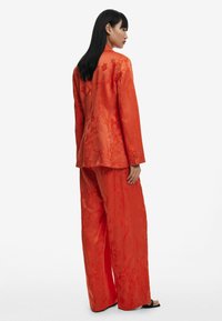 Rode zijden pak met een textuur van een bloemmotief, een op maat gemaakte blazer met een enkele knoop, en broek met wijde pijpen en een gladde afwerking.