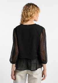 Sort bluse med teksturerede, gennemsigtige lange ærmer og peplumnederdel. Stoffet har et prikket mønster og en glat ryg.