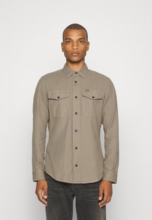 Hombre con una camisa beige abotonada y con bolsillos en el pecho, de pie frente a un fondo blanco liso.