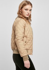 Jeune femme aux cheveux blonds ondulés portant une veste matelassée beige et un pantalon noir, debout de profil sur un fond uni.