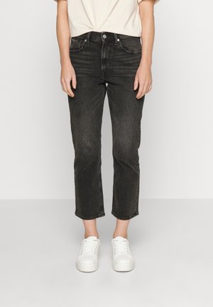 Person trägt schwarze, gerade geschnittene Jeans mit schmalem Bein und weiße Plateausneaker, steht vor hellem Hintergrund.