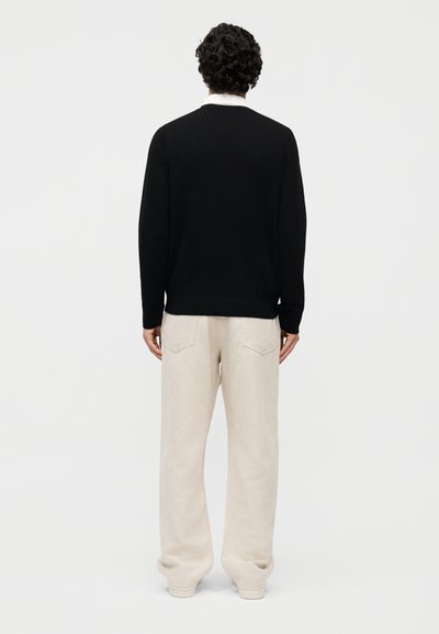 Άνδρας με σγουρά μαλλιά, φορώντας μαύρο πουλόβερ με μακριά μανίκια και χαλαρό μπεζ παντελόνι, στέκεται με την πλάτη στη λευκή, μονόχρωμη επιφάνεια.