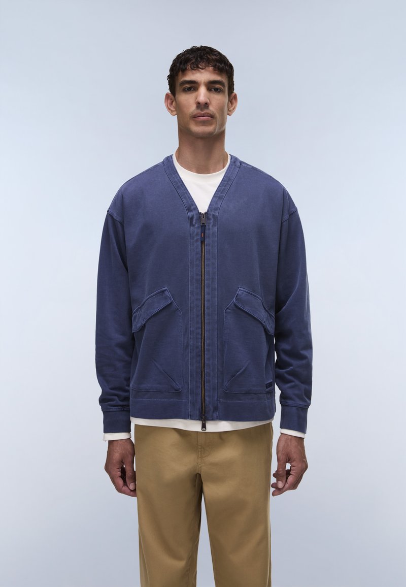 Μπλε μπουφάν με φερμουάρ, κατασκευασμένο από βαμβάκι, με V-λαιμόκοψη, δύο εμπρόσθιες τσέπες και άνετη εφαρμογή. Συνδυάζεται με ανοιχτόχρωμο παντελόνι.