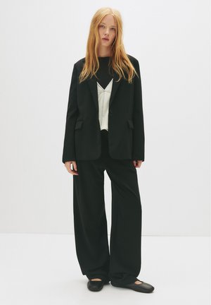 Mladá žena s dlhými červenými vlasmi, oblečená v čiernom obleku so širokými nohavicami, bielym topom a čiernymi plochými topánkami na jednoduchom bielom pozadí.