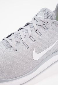 Jasnoszary but sportowy z teksturowaną siateczką na górze, płaskimi sznurowadłami oraz białym logo swoosh z boku; posiada amortyzowaną podeszwę.