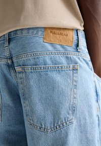 Vaqueros de mezclilla azul claro con acabado texturizado, que presentan un parche de cuero marrón con la etiqueta "PULL&BEAR" en la cintura y bolsillos traseros estándar.