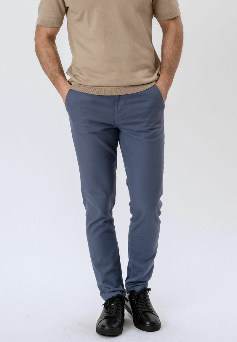 Homme portant une chemise beige à manches courtes, un pantalon slim bleu et des baskets noires, debout les mains dans les poches devant un fond uni.