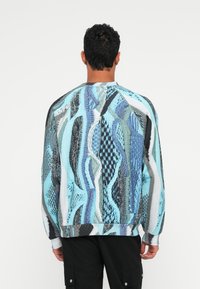 Stickad tröja i blå, svart och grå mönster; har en texturerad tyg med rund hals och ribbade muddar. Bakifrån visas intrikata designdetaljer.