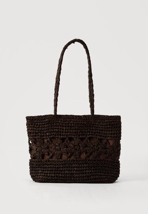 Geantă tote maro închis împletită cu modele florale și două mânere lungi împletite pe un fundal simplu, deschis la culoare.