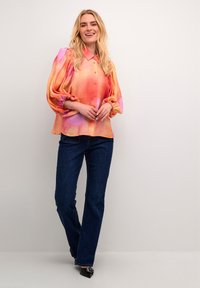 Bluză colorată, ușoară, în nuanțe gradient de portocaliu și roz, cu închidere pe butoane și mâneci bufante, asortată cu blugi de culoare albastru închis.