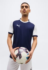 Jeune homme vêtu d'une tenue de sport Puma bleu marine et blanche tenant un ballon de football blanc avec des motifs noirs et roses sur un fond uni.