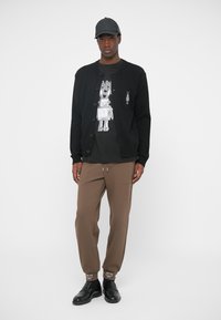 Cárdigan negro con botones sobre una camiseta gráfica, combinado con pantalones marrones ajustados y zapatos negros. El conjunto presenta texturas y colores distintos.