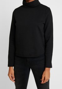Maglia a collo alto nera realizzata in un tessuto morbido e elasticizzato, con maniche lunghe e una vestibilità leggermente cropped, caratterizzata da una texture liscia.