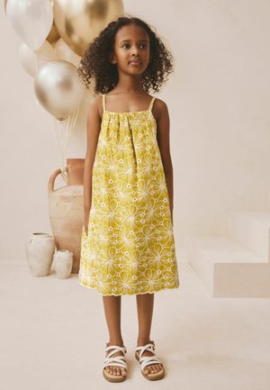 Mladé dievča v žltých kvetovaných šatách bez rukávov a bielych sandáloch stojí pri zlatých a bielych balónikoch a keramických váza na svetlom pozadí.