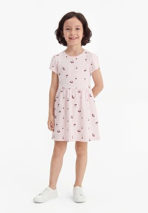 Νεαρή κοπέλα που χαμογελά, φορά ροζ παστέλ κοντομάνικο φόρεμα με σχέδιο κερασιών και λευκά αθλητικά παπούτσια, στέκεται μπροστά σε απλό λευκό φόντο.