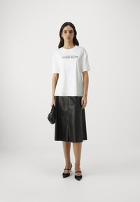 T-shirt blanc à manches courtes avec le logo "GUESS JEANS", associé à une jupe midi en cuir noir et des chaussures à talons hauts noires. Silhouette équilibrée.