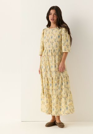 Kvinde står iført en gul blomsterprint lang kjole med trekvartærmer og brune flade sko mod en ensfarvet off-white baggrund.