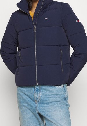 Chaqueta acolchada azul marino con cremallera frontal y bolsillos con cremallera, usada con vaqueros azul claro, logotipo de la marca en el pecho y la manga.